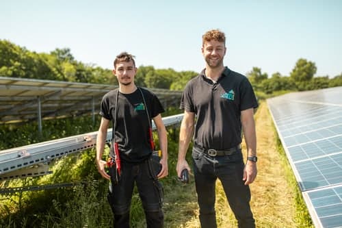 Zwei Männer in schwarzen T-Shirts mit Logo stehen zwischen Reihen von Solarmodulen unter blauem Himmel.