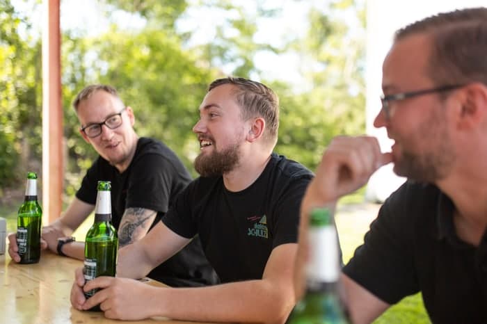 Drei Männer sitzen an einem Holztisch mit grünen Bierflaschen und unterhalten sich lächelnd im Freien.
