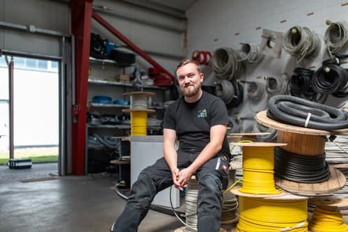 Junger Mann mit Bart sitzt in einer Werkstatt voller Kabelrollen, trägt ein schwarzes T-Shirt und Arbeitshosen.