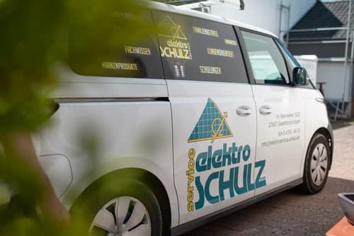 Weißer Elektro Schulz Service Transporter mit Logo und Aufschriften, teilweise von Blättern verdeckt.