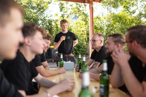 Ein Mann steht lächelnd vor einer Gruppe von sechs sitzenden Männern an einem Holztisch mit Bierflaschen.