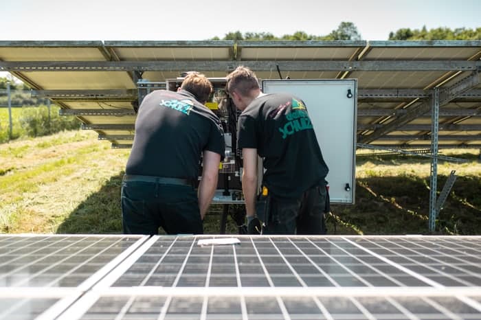 Zwei Techniker in schwarzen Hemden mit „SCHULZ“-Logo arbeiten an einem Wechselrichter unter Solarmodulen.