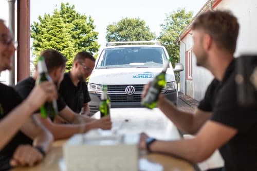 Vier Männer sitzen an einem Tisch mit Bierflaschen, im Hintergrund ein weißer VW-Bus mit "Schulz"-Logo.