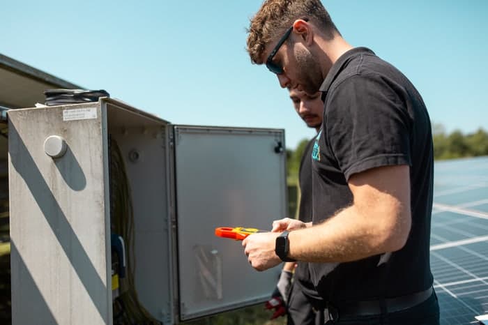 Zwei Männer prüfen eine Elektrobox mit Messgerät neben Solarmodulen unter blauem Himmel.
