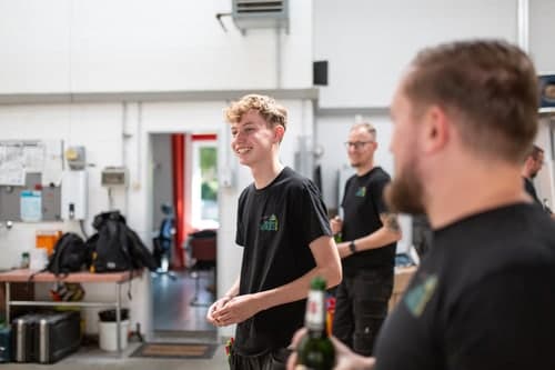 Ein junger Mann mit blonden Locken in einem schwarzen T-Shirt lacht in einem hellen Lagerhaus, während ein anderer Mann im Vordergrund eine Flasche hält.